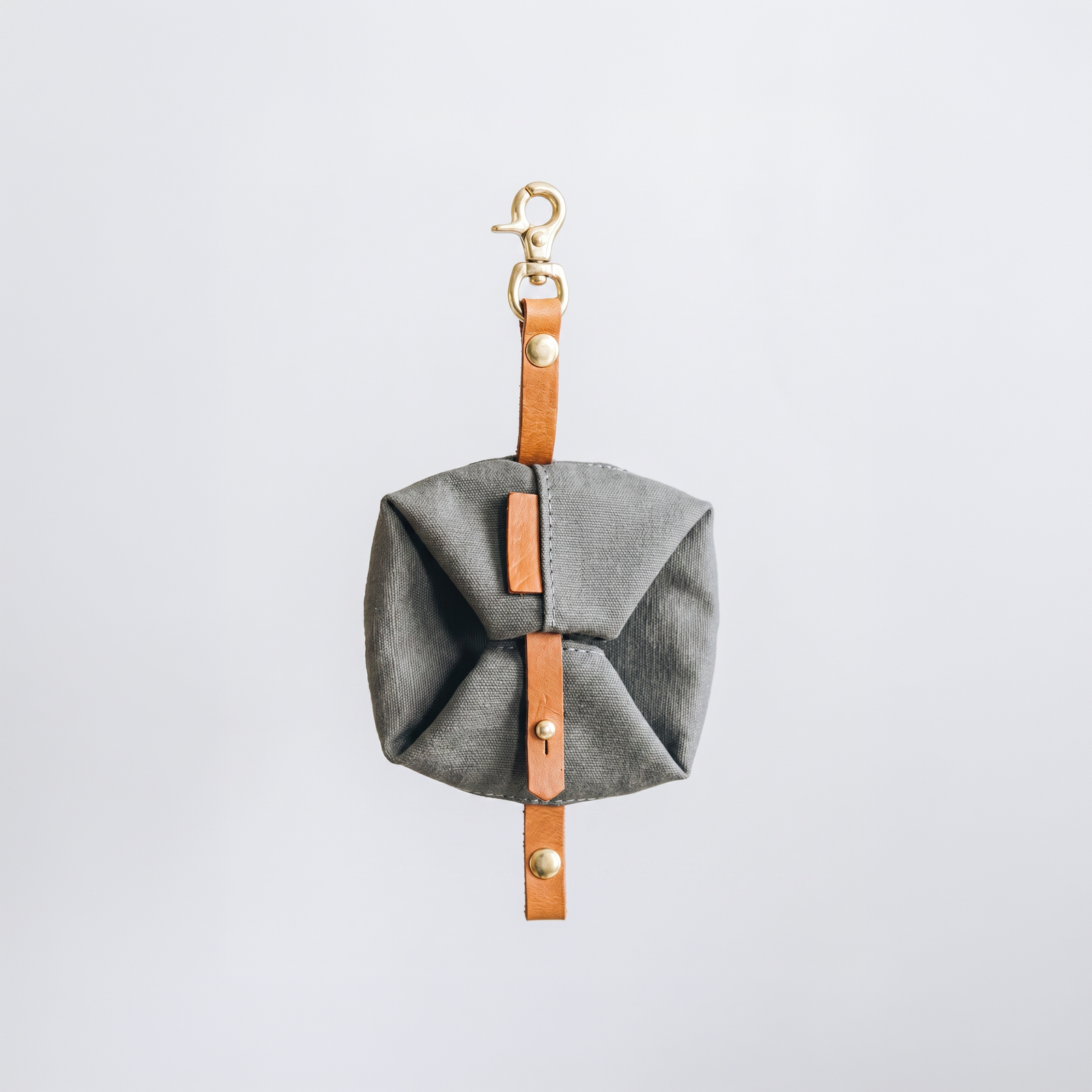 Gamelle pliable pour chien en tissu de couleur gris foncé, repliée en pochette de transport compacte avec sangle de fermeture en cuir marron, bouton pression et mousqueton en laiton doré sur fond blanc.